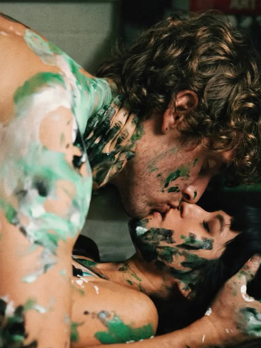 A couple kissing, covered in green paint, while making a painting with their bodies on canvas