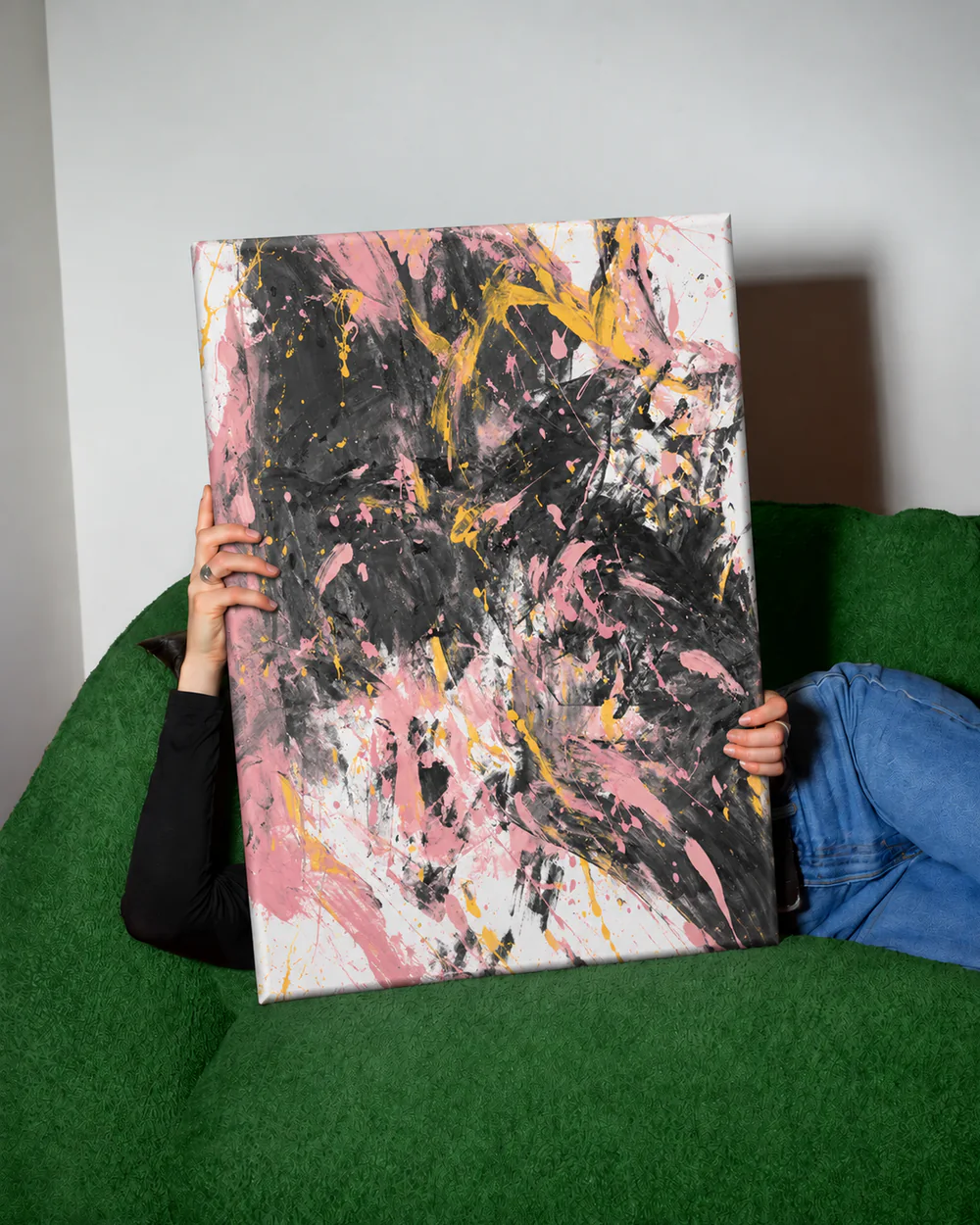 A person on a green sofa holds up a pink and black painting on a 120 × 90 cm frame