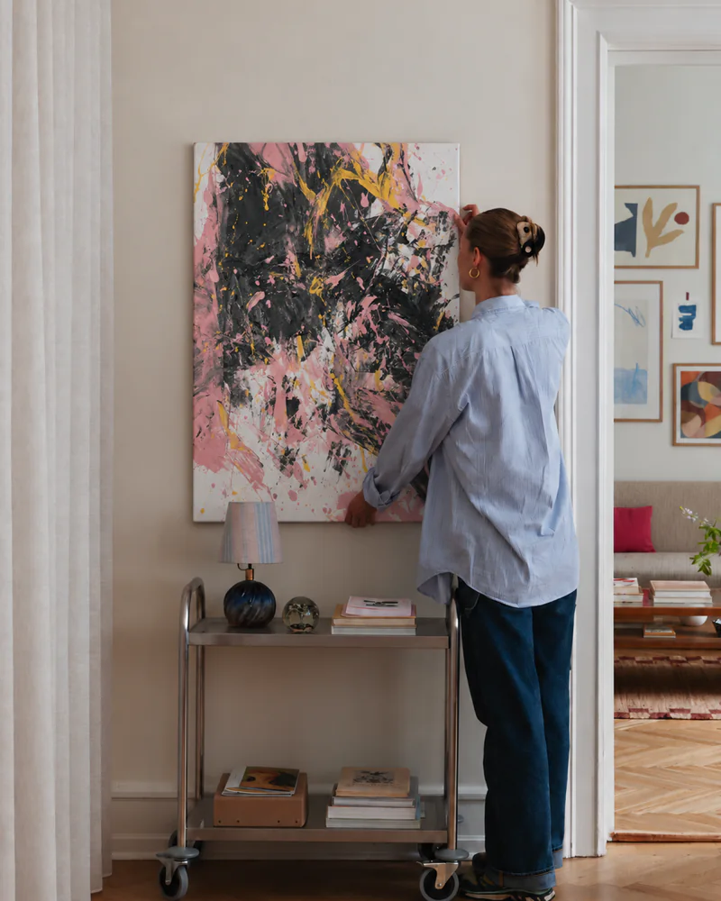 The same pink and black painting being hung on a wall above a metal trolley with a lamp