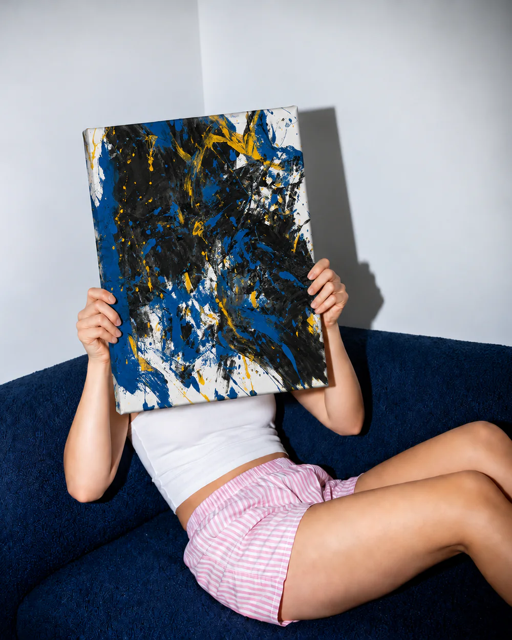 A person on a navy sofa holds a blue and black painting on a 50 × 40 cm frame in front of her face