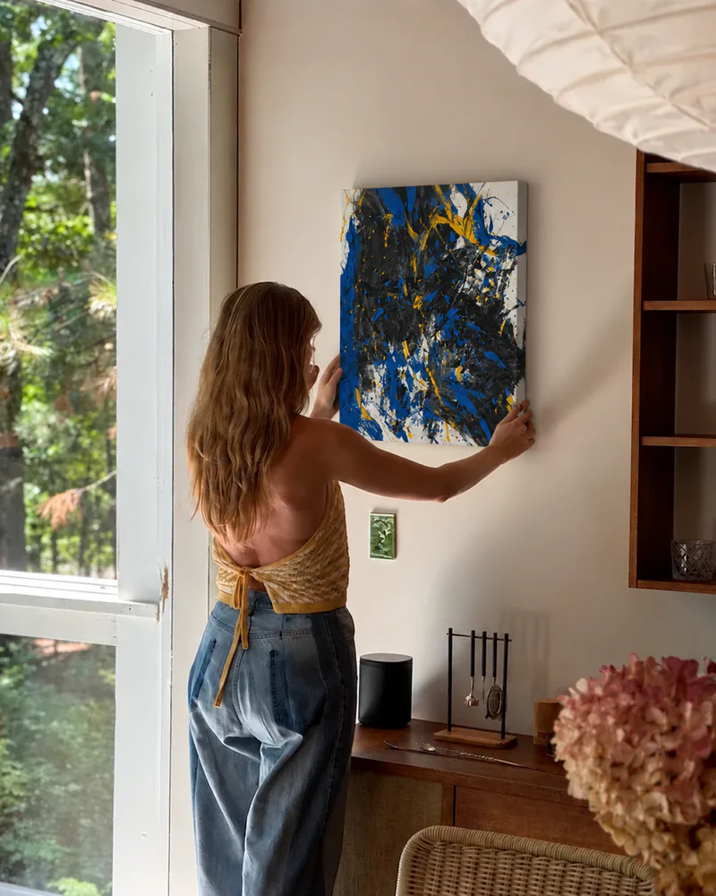 The same blue and black painting being hung on a wall by a window, next to wooden shelves
